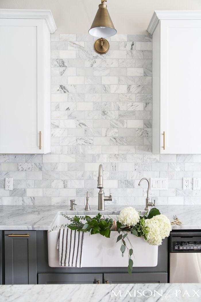 Two-toned gray and white cabinets, marble subway tile, Carrara countertops, a big farmhouse sink, and brass hardware give this kitchen a classic yet modern look.