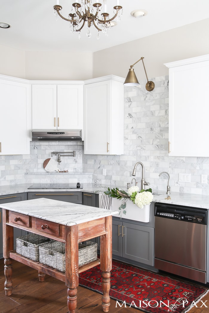 Two-toned gray and white cabinets, marble subway tile, Carrara countertops, a big farmhouse sink, and brass hardware give this kitchen a classic yet modern look.