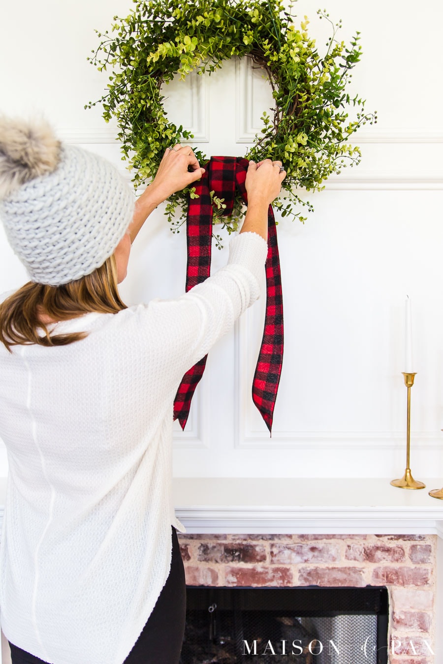 woman hanging red buffalo check ribbon on green wreath over mantel | Maison de Pax
