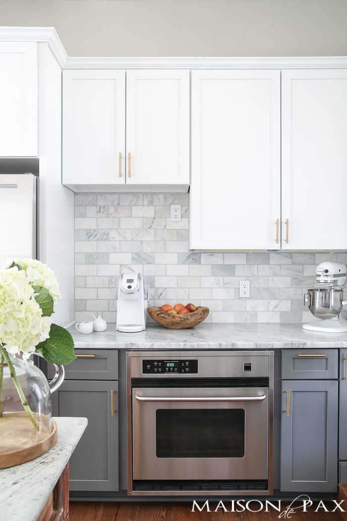 Two-toned gray and white cabinets, marble subway tile, Carrara countertops, a big farmhouse sink, and brass hardware give this kitchen a classic yet modern look.