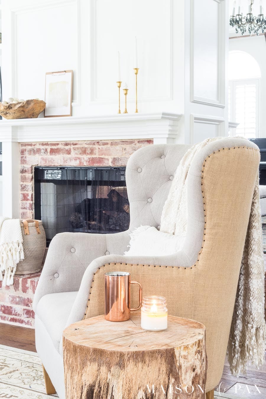 tufted armchair with log side table before a firplace with antique brick and white molding, all accented with gold and copper