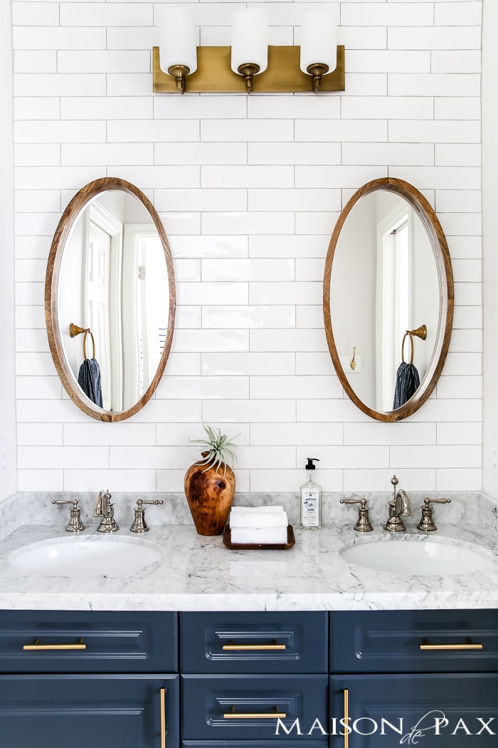 wood mirrors hung on white tile wall above navy vanity in bath