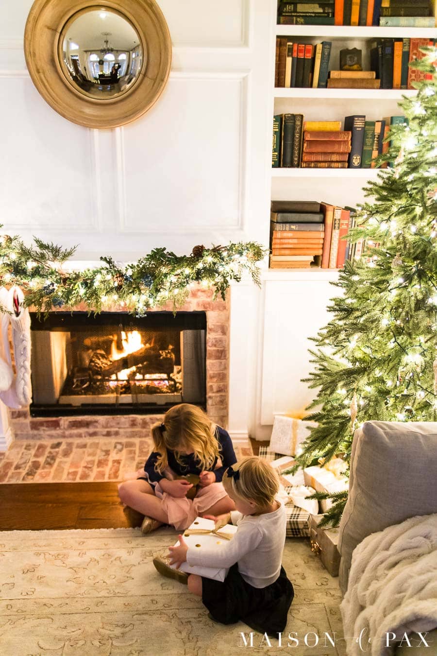 girls looking at presents under Christmas tree by fireplace | Maison de Pax